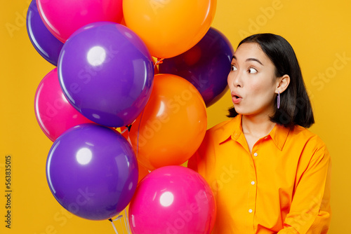 Close up happy fun surprised young woman wearing casual clothes celebrating holding looking at bunch of colorful air balloons isolated on plain yellow background. Birthday 8 14 holiday party concept.