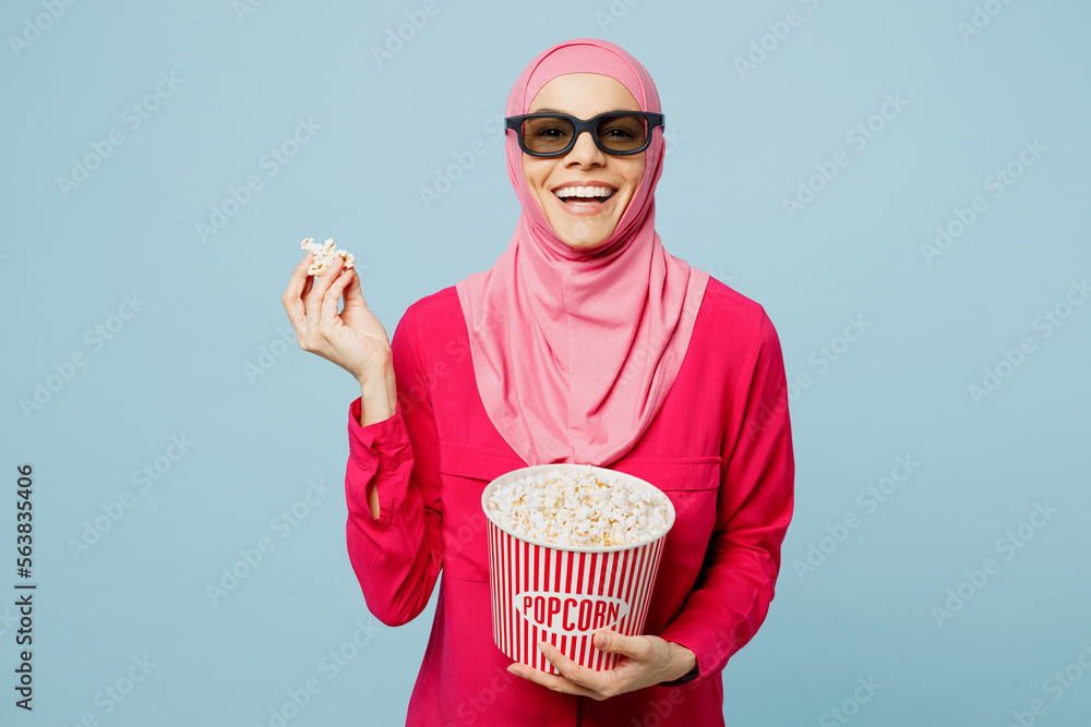 Young laughing happy arabian muslim woman woman in 3d glasses abaya hijab watch comedy movie film hold bucket of popcorn in cinema look camera isolated on plain blue cyan background studio portrait.