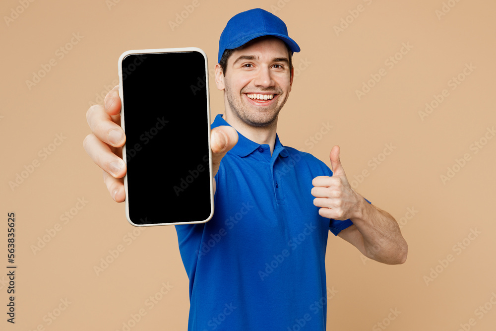 Delivery guy employee man wearing blue cap t-shirt uniform workwear work as dealer courier hold in hand use mobile cell phone with blank screen area show thumb up isolated on plain beige background. - obrazy, fototapety, plakaty 