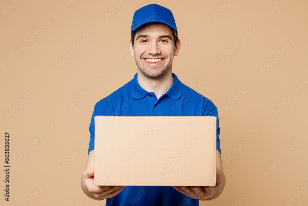 Professional smiling cheerful fun delivery guy employee man wear blue cap t-shirt uniform workwear work as dealer courier hold cardboard box isolated on plain light beige background. Service concept - obrazy, fototapety, plakaty 