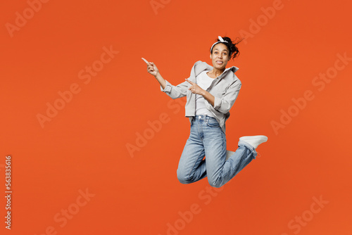 Full body young woman of African American ethnicity wears grey shirt headband jump high point index finger aside on area isolated on plain orange background studio portrait. People lifestyle concept.