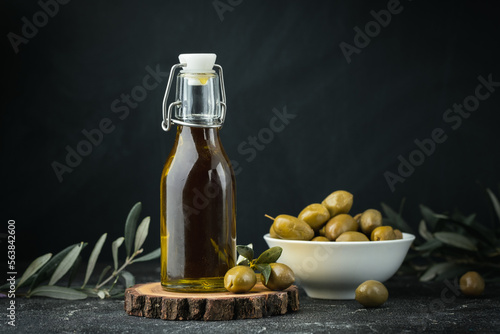 Green olives in a white bowl next to a bottle with olive oil and leaves on a black background. Bottle of cold pressed oil. Traditional Greek and Italian food.