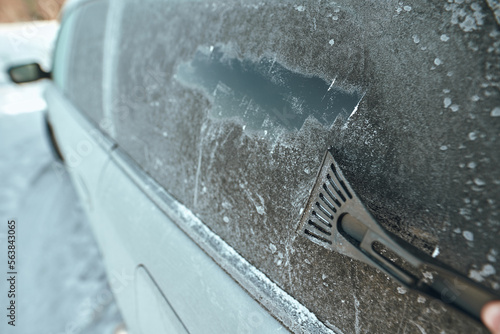 Man is cleaning an icy window on a car with an ice scraper. Cold snowy and frosty morning. Focus on the ice scraper.