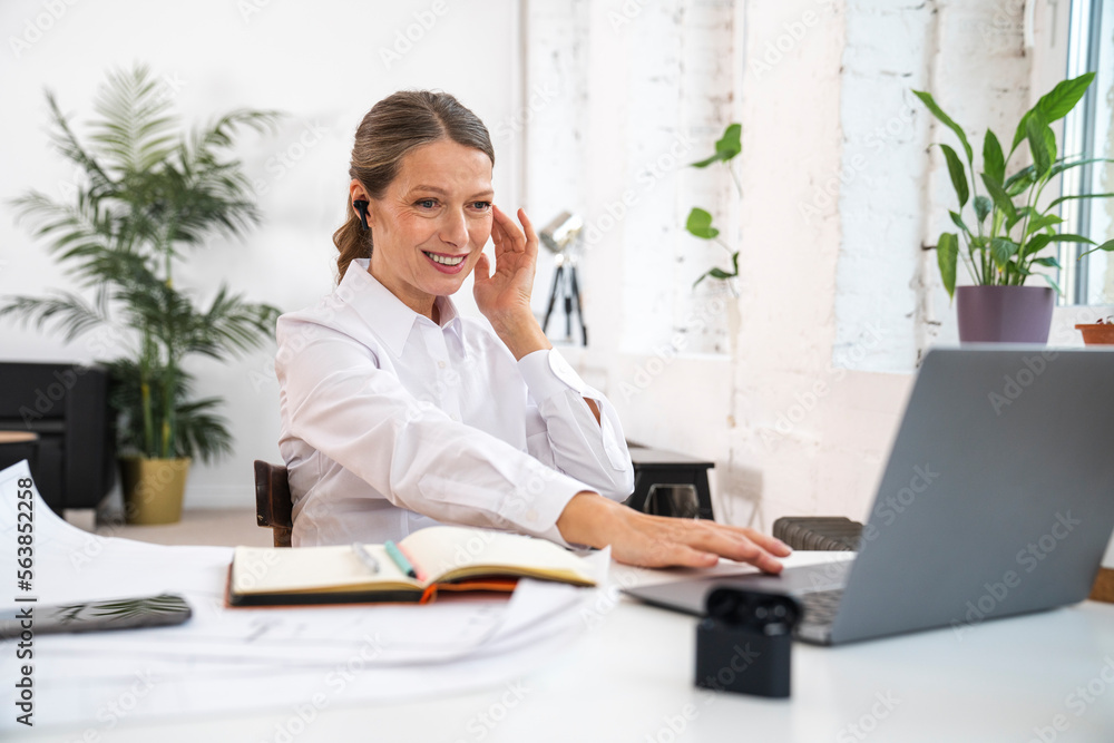 Smiling businesswoman wearing wireless in-ear headphones using laptop in office Smiling businesswoman wearing wireless in-ear headphones using laptop in office