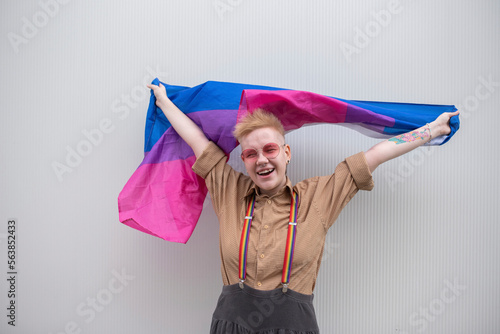 Happy non-binary person holding bisexual flag in front of white wall photo