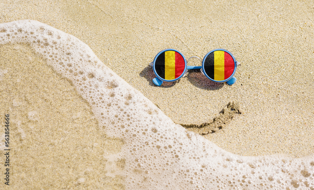 Obraz premium Sunglasses with flag of the Belgium on a sandy beach. Nearby is a sea lightning and a painted smile.