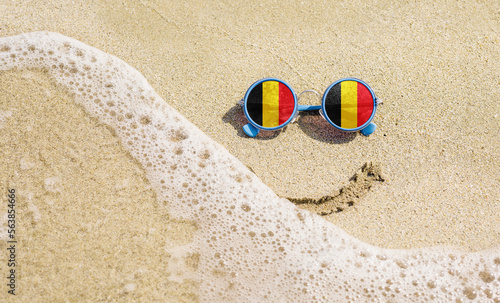 Sunglasses with flag of the Belgium on a sandy beach. Nearby is a sea lightning and a painted smile.