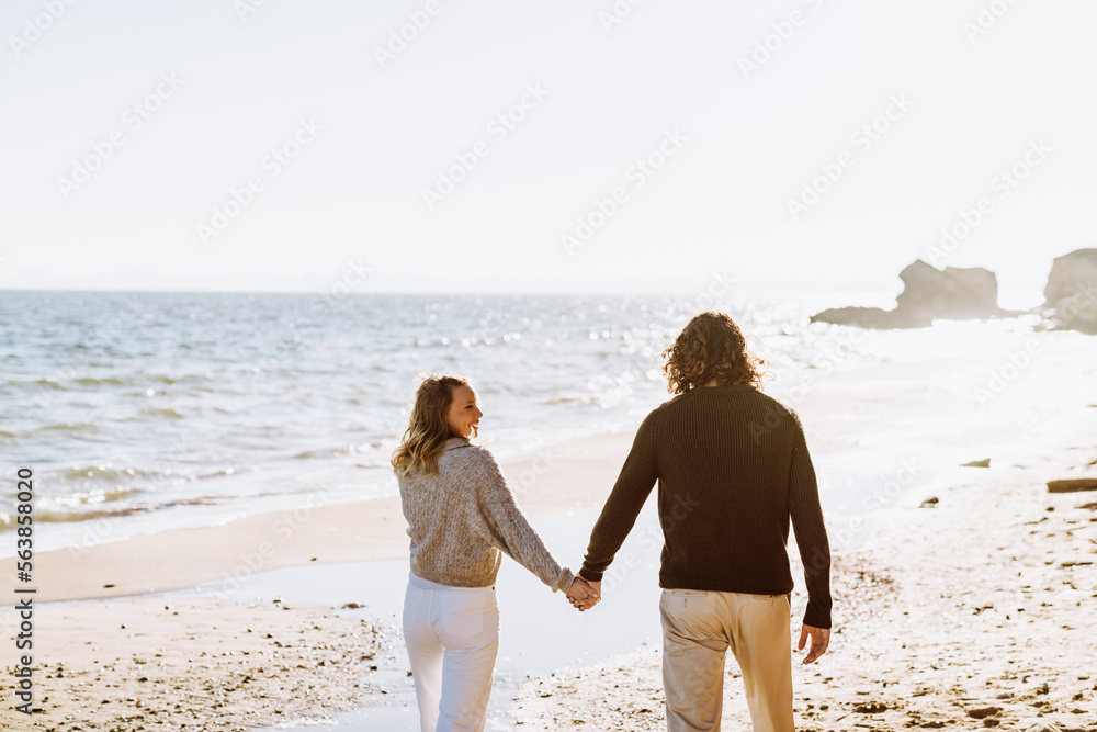 Young smiling couple holding hands during walk at sunny beach