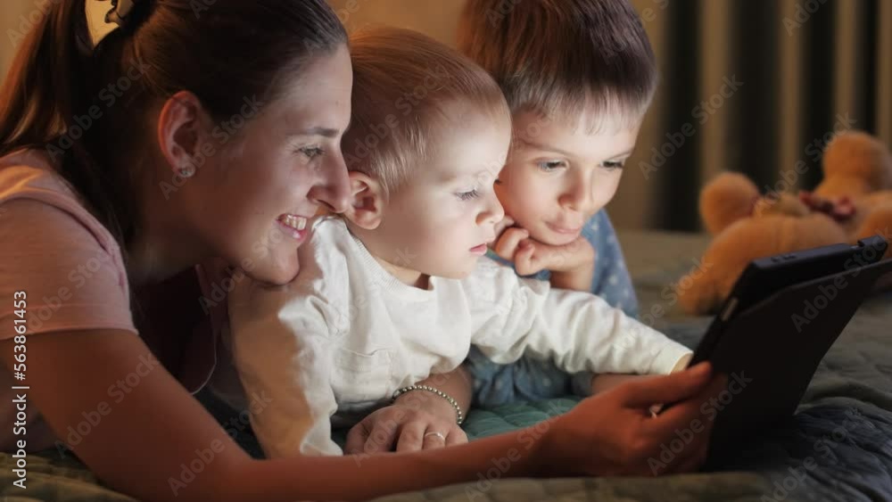 Two smiling brothers in pajamas playing on tablet computer with mother before going to sleep. Family having time together, parenting, happy childhood and entertainment Two smiling brothers in pajamas playing on tablet computer with mother before going to sleep. Family having time together, parenting, happy childhood and entertainment