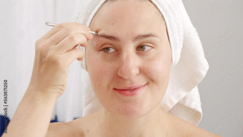 Closeup of smiling woman looking in mirror and tweezing eyebrows with tweezers at the bathroom. Concept of beautiful female, makeup at home, skin care and domestic beauty industry Closeup of smiling woman looking in mirror and tweezing eyebrows with tweezers at the bathroom. Concept of beautiful female, makeup at home, skin care and domestic beauty industry