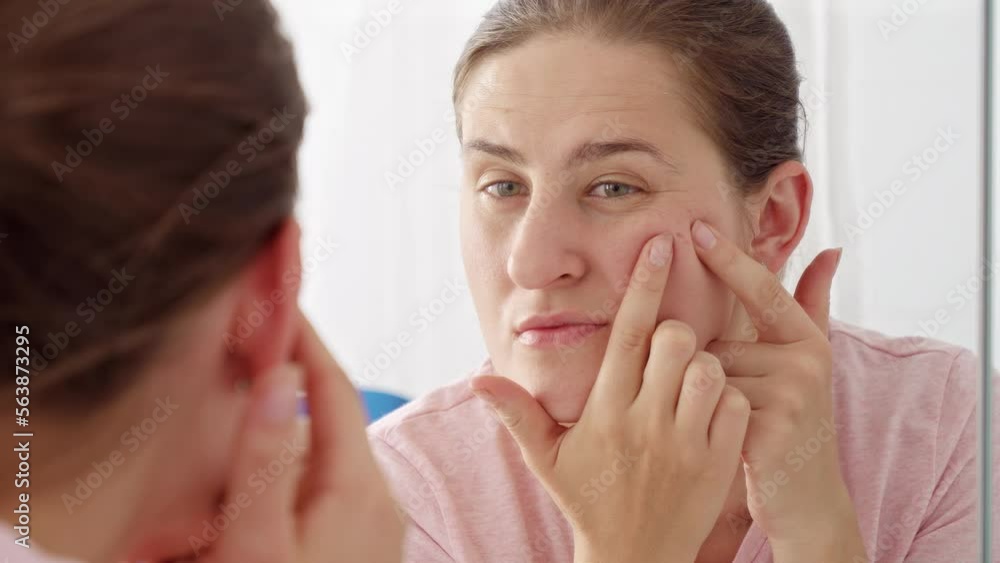 Young woman looking at her reflection and squeezing pimples and acne. Concept of female aging, skin issues, facial treatment and care Young woman looking at her reflection and squeezing pimples and acne. Concept of female aging, skin issues, facial treatment and care