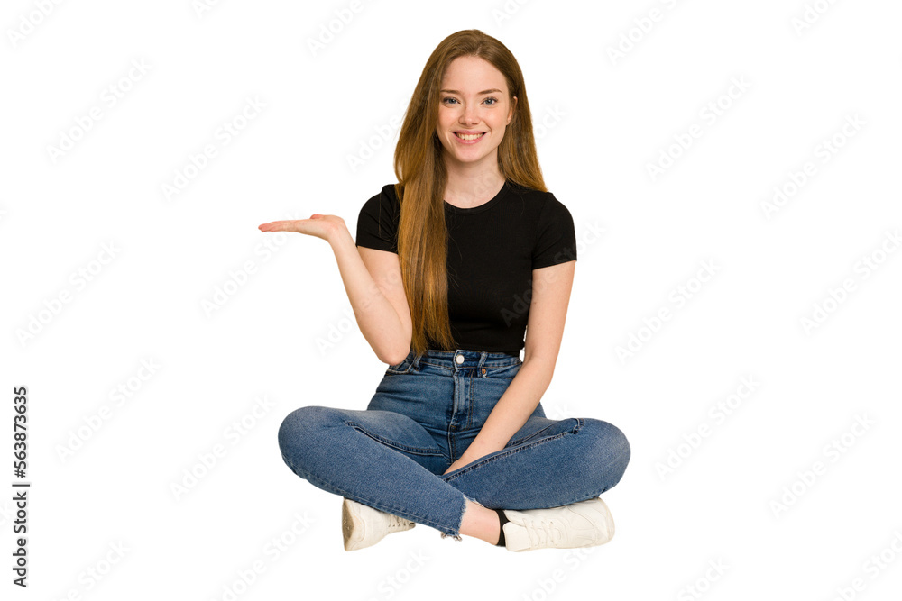 Young redhead woman sitting on the floor cut out isolated showing a copy space on a palm and holding another hand on waist.