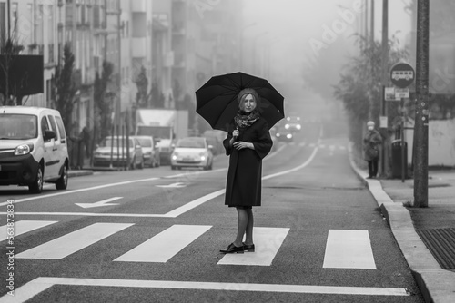 A woman with an umbrella stands in the street in cloudy weather. Black and white photo.