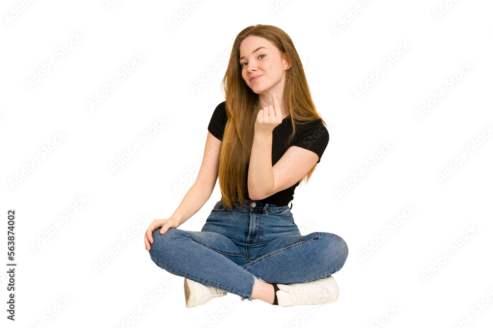 Young redhead woman sitting on the floor cut out isolated pointing with finger at you as if inviting come closer.