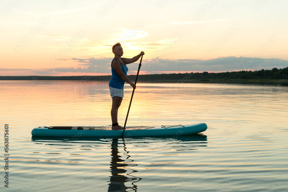 A woman in shorts and a top on a SUP board with a paddle at sunset swims in the water of the lake in the glare of the setting sun.