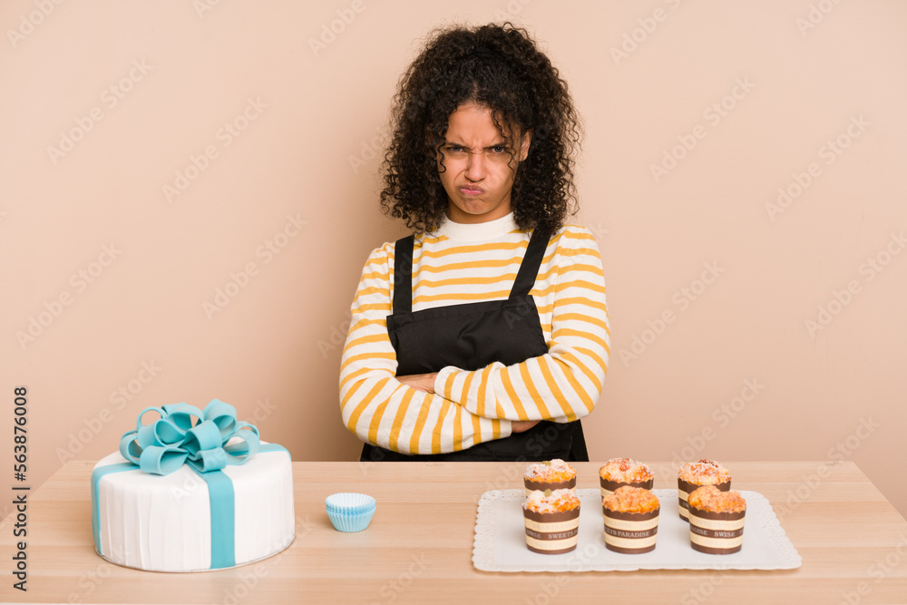 Young african american woman preparing a sweet cake and muffins on a table unhappy looking in camera with sarcastic expression. Young african american woman preparing a sweet cake and muffins on a table unhappy looking in camera with sarcastic expression.