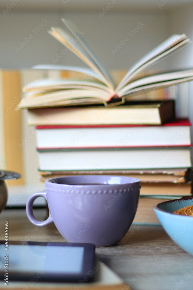 Stack of books, e-reader, reading glasses, bowl of biscuits, apples and cup of tea on the table. Bookshelf in the background. Selective focus. Stack of books, e-reader, reading glasses, bowl of biscuits, apples and cup of tea on the table. Bookshelf in the background. Selective focus.