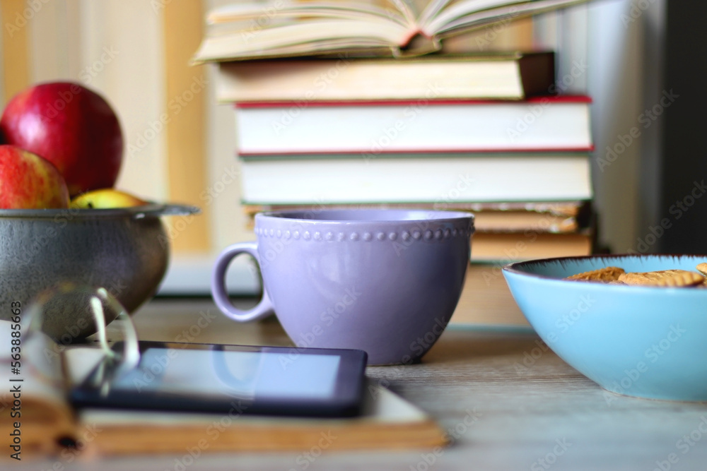 Stack of books, e-reader, reading glasses, bowl of biscuits, apples and cup of tea on the table. Bookshelf in the background. Selective focus. Stack of books, e-reader, reading glasses, bowl of biscuits, apples and cup of tea on the table. Bookshelf in the background. Selective focus.