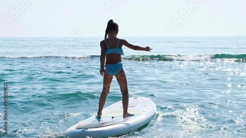 Girl learns with pleasure to keep balance on SUPboard against background of blue sea water and setting sun.