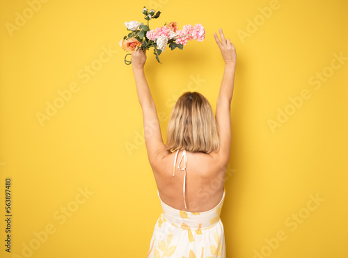 Fotografía Back view Image of a beautiful amazing young blonde woman posing isolated over yellow wall background holding flowers
