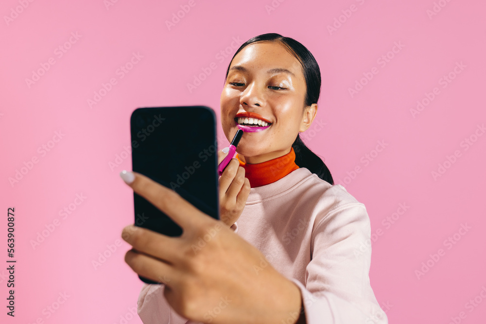 Gen Z female using a smartphone to apply lipstick in a studio Gen Z female using a smartphone to apply lipstick in a studio