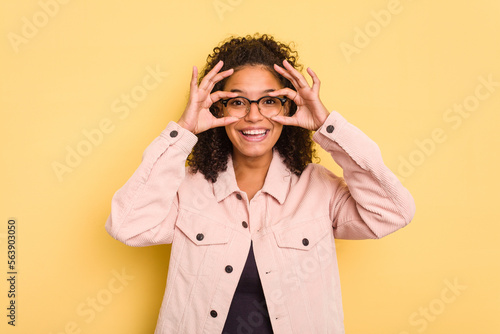Young Brazilian curly hair cute woman isolated on yellow background showing okay sign over eyes