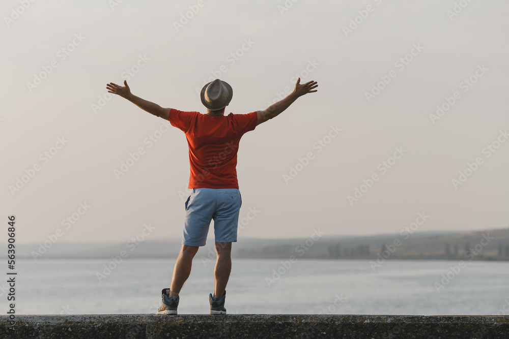 Young hipster man enjoying the picturesque view of Italian lake with raised hands in hat, red t-shirt, blue shorts Young hipster man enjoying the picturesque view of Italian lake with raised hands in hat, red t-shirt, blue shorts