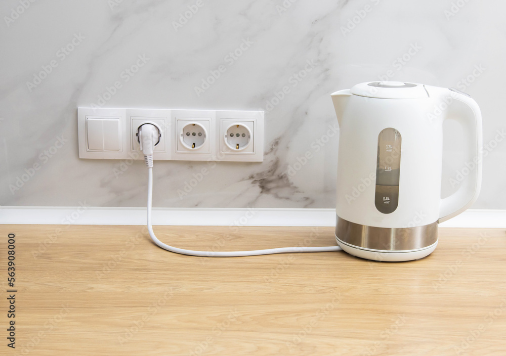A white modern electric kettle stands on a table against the background of sockets. Boiling water. Electricity consumption by household electrical appliances. Electricity price. Copy space for text A white modern electric kettle stands on a table against the background of sockets. Boiling water. Electricity consumption by household electrical appliances. Electricity price. Copy space for text