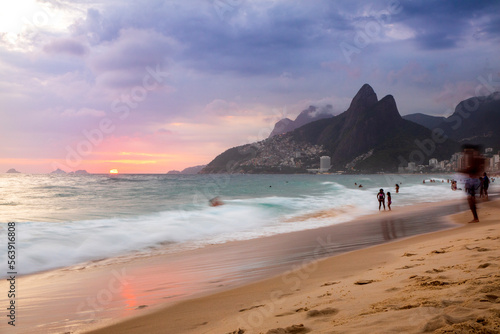 Long exposure of beachgoers in Rio de Janeiro, Brazil playing on the sand and in the water of the Atlantic Ocean as the sun sets over the sea near Ipanema Beach.