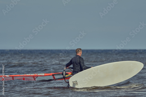 A mature man in a hydrosuit walks through shallow water and carries a sailboard on a sunny autumn day.