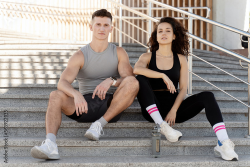 Beautiful fitness couple wearing stylish sportive clothes sitting at the stairs and posing on city street