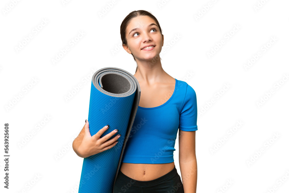 Teenager caucasian girl going to yoga classes while holding a mat over isolated background looking up while smiling
