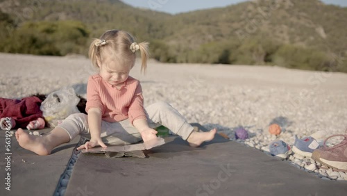 Toddler sits on a camping mat, trying to deal with camping equipment. Tiddler sits on a pebble beach photo