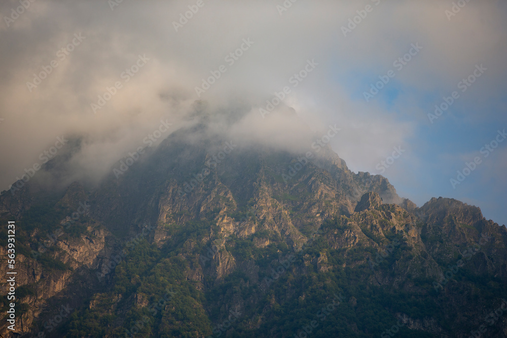 Dark atmospheric surreal landscape with a dark rocky mountain peak in low clouds in a gray cloudy sky. A gray low cloud on a high peak. High black rock in low clouds. Surreal gloomy mountains. Dark atmospheric surreal landscape with a dark rocky mountain peak in low clouds in a gray cloudy sky. A gray low cloud on a high peak. High black rock in low clouds. Surreal gloomy mountains.