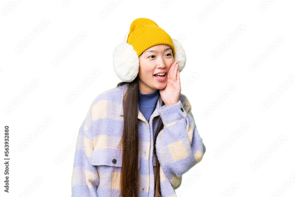 Young Asian woman wearing winter muffs over isolated chroma key background shouting with mouth wide open to the lateral Young Asian woman wearing winter muffs over isolated chroma key background shouting with mouth wide open to the lateral