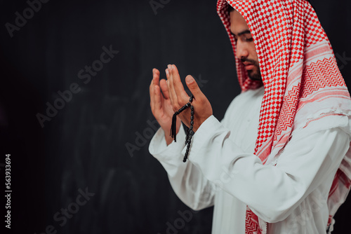 Arabian man in traditional clothes making traditional prayer to God, keeps hands in praying gesture in front of black chalkboard representing modern islam fashion and ramadan kareem concept