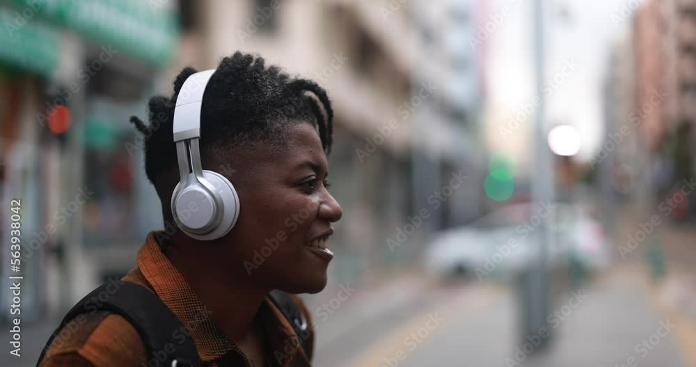 Young african woman waiting at bus station while listening music playlist with wireless headphones Young african woman waiting at bus station while listening music playlist with wireless headphones