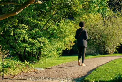 Black woman walks in a park. Unrecognizable woman with afro hair strolls along a tree-lined avenue during sunset. Image of serenity on an autumn day. Back view of womanin black dress with shoulder bag photo