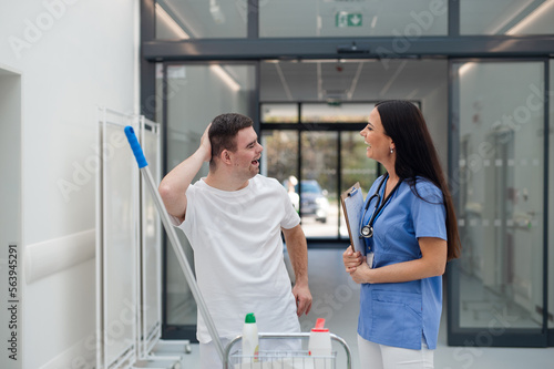 Young man with down syndrome working in a hospital as cleaner, talking to nurse, having fun. Concpet of integration people with disability into society.
