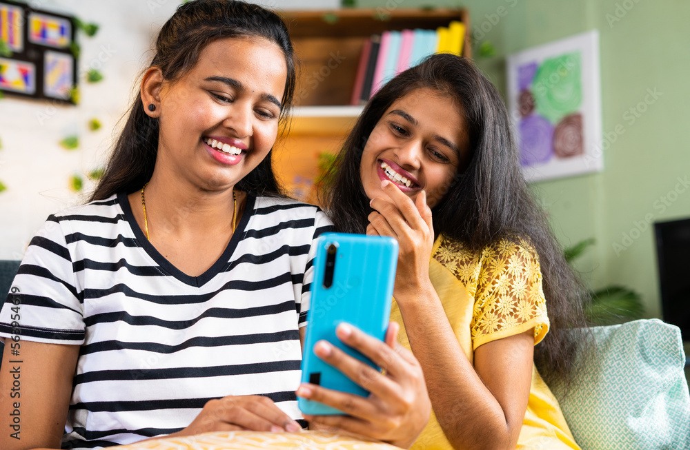 laughing sibling girls while using mobile phone on sofa at home - concept of entertainment, social media and internet connection. laughing sibling girls while using mobile phone on sofa at home - concept of entertainment, social media and internet connection.