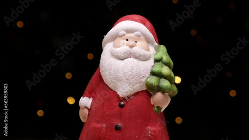 Ceramic Santa Claus figurine. Festive toy decoration for Christmas. Tree flickering lights bokeh in background. Xmas Holiday Season winter composition.