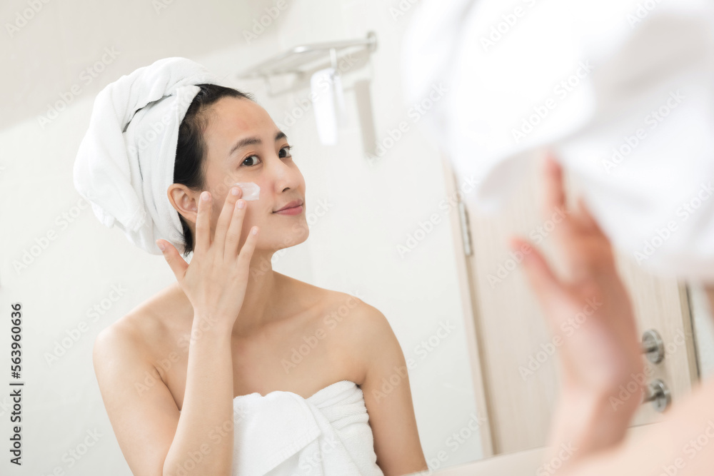 Happy young Asian woman applying face lotions while wearing a towel and touching her face in bathroom Happy young Asian woman applying face lotions while wearing a towel and touching her face in bathroom
