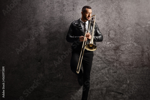 Man in a leather jacket holding a trombone and standing against a gray wall
