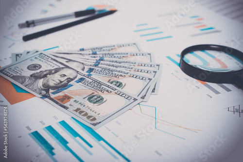 Close-up of money, business documents, magnifier, pen and pencil in workplace. Low angle view, selective focus on one hundred dollar bills