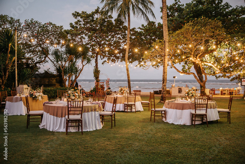 Wedding banquet or gala dinner decorated with garlands. Round tables styled with flowers, candles and accessories. Open air festive banquet on green lawn with sea view during dusk