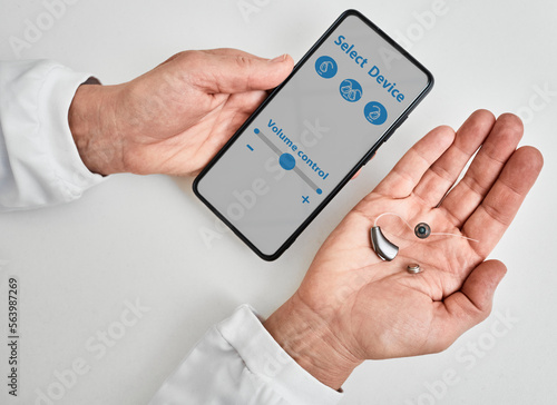 Doctor audiologist showing smartphone app for adjusting hearing aid holding smartphone in one hand and BTE hearing aid in other. Manage hearing aid settings via smartphone