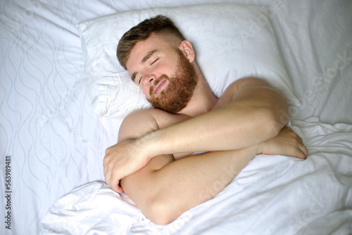 Handsome young calm sleepy bearded man with beard is sleeping well on side in bed in bedroom on pillow with his eyes closed in the morning  resting. Good healthy dream  rest. White linens. Top view.