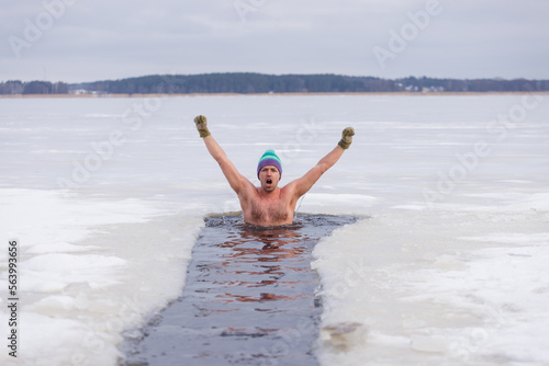 Winter swimming. Man in frozen lake ice hole. Swimmers wellness in icy water. How to swim in cold water. Beautiful young man in zen meditation. Hat and gloves swimming clothes. Nature lake energy