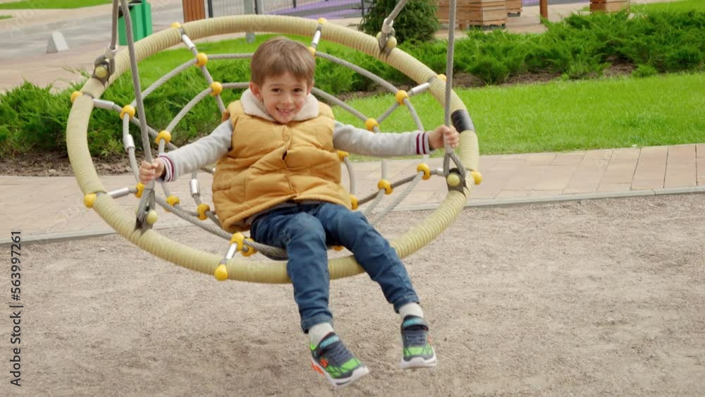 Cheerful boy sitting in wicker rope swing and riding up high. Active child, sports and development, kids playing outdoors. Cheerful boy sitting in wicker rope swing and riding up high. Active child, sports and development, kids playing outdoors.