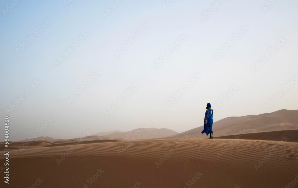Woman standing on a large sand dune in the sahara. Merzouga desert. Morocco. One person standing alone in the desert looking towards the dunes. 